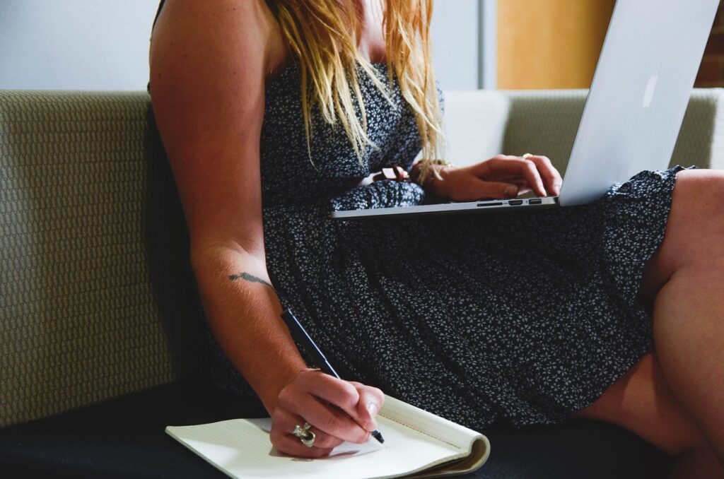 woman sitting on a couch meal planning with one hand on a laptop and the other writing something in a notebook